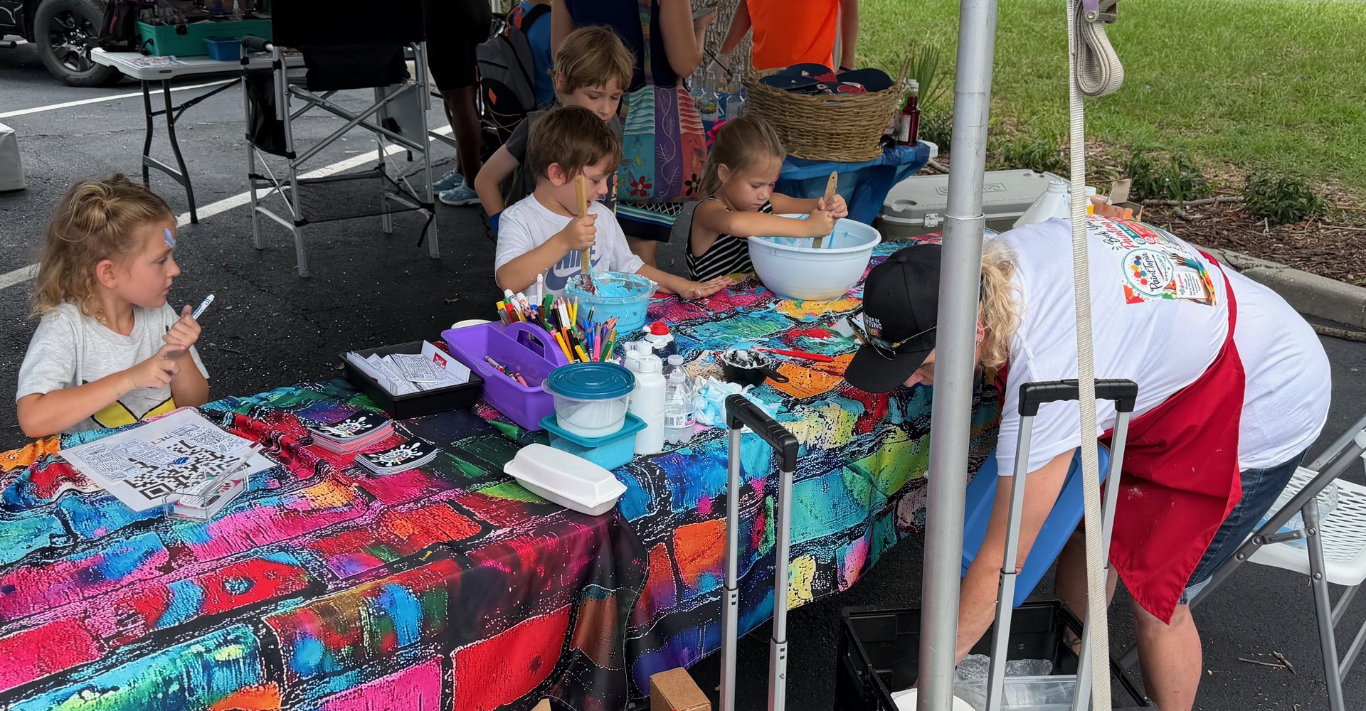 Children and an adult participating in outdoor art activity at a colorful covered table with art supplies, brushes, and paint.