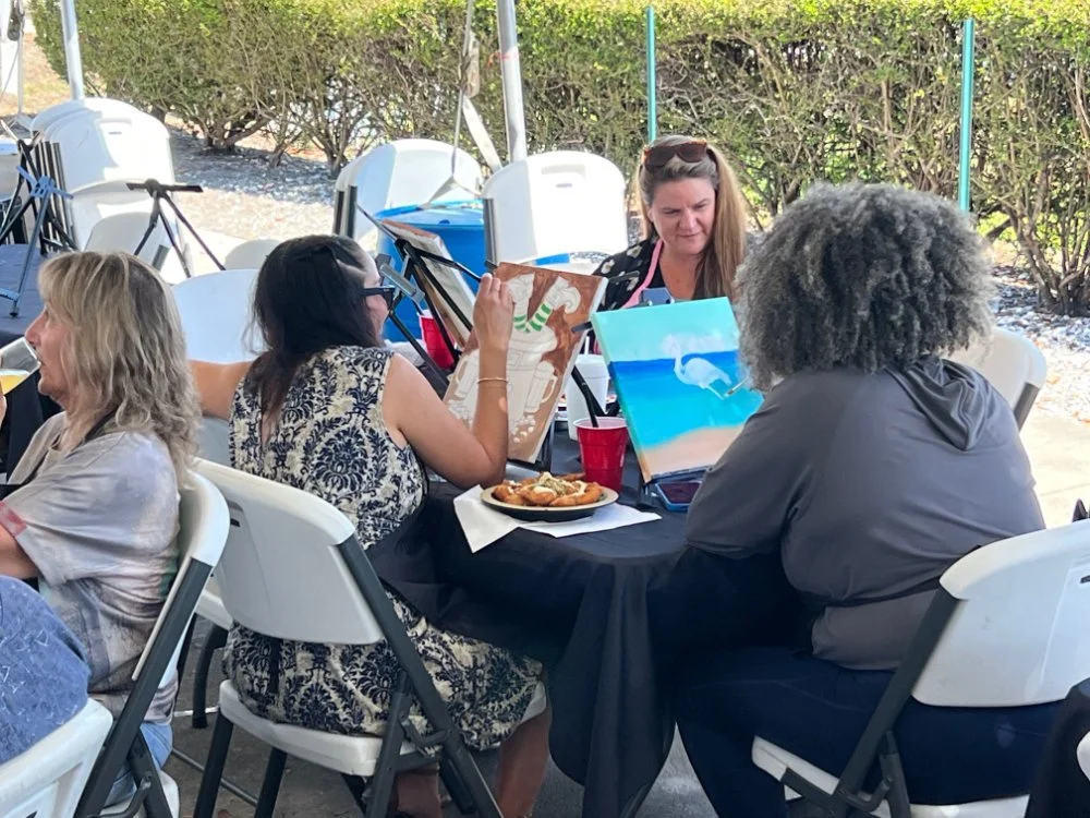 Group of women sitting around a table, painting on canvases, with snacks and drinks, outdoors in a garden-like setting.