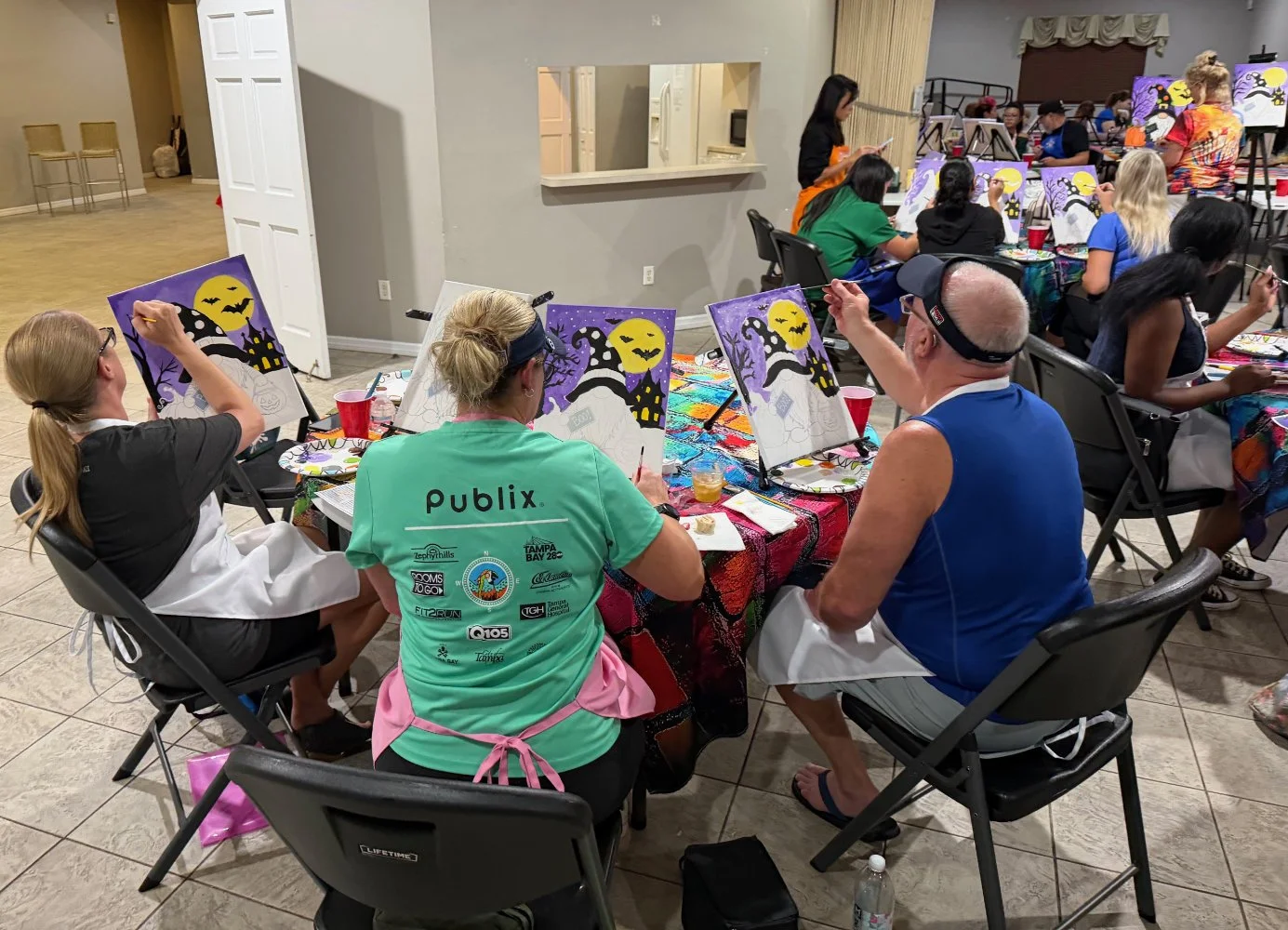 People painting Halloween-themed pictures at a group art event with children and adults seated at tables.