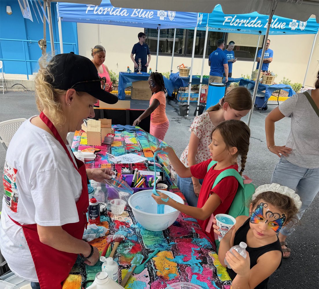 Children at a face painting booth with one girl already painted like a butterfly, and other kids getting their faces painted at a booth under tents labeled 'Florida Blue' at an outdoor event.