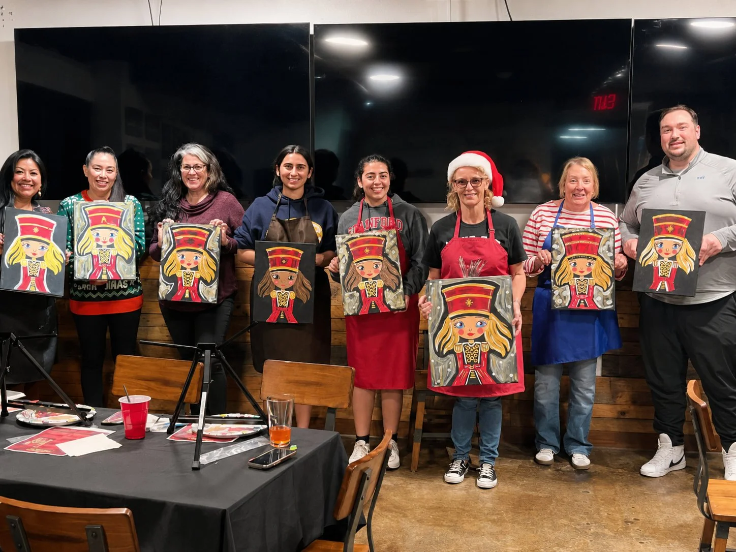 Group of eight women and one man standing indoors, holding paintings of a girl in a red dress and tall red hat, some wearing festive Christmas attire including a Santa hat and red apron, smiling and facing the camera