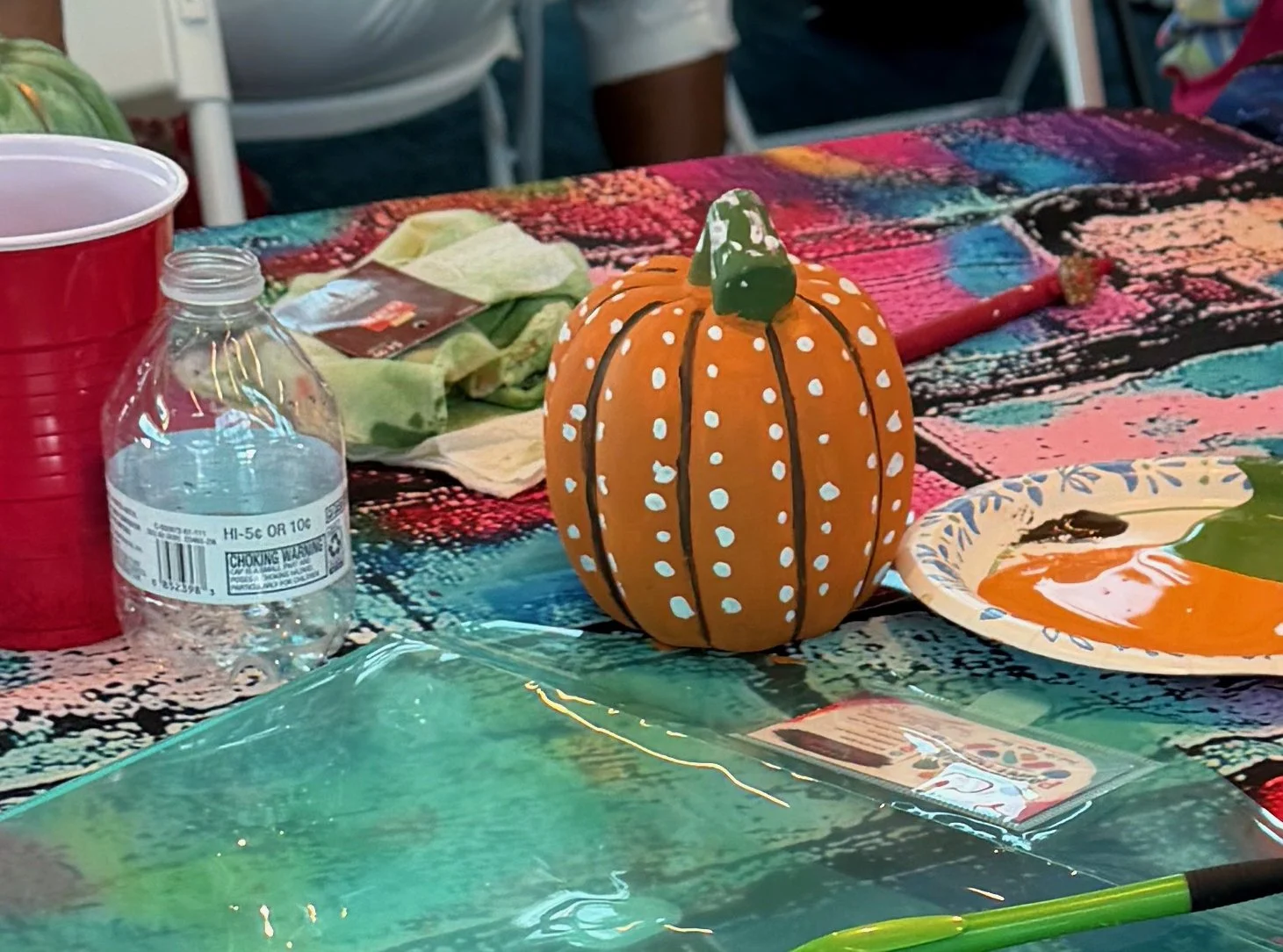A painted orange pumpkin with white dots and black lines, and a green top, sitting on a colorful tablecloth surrounded by art supplies and cups.