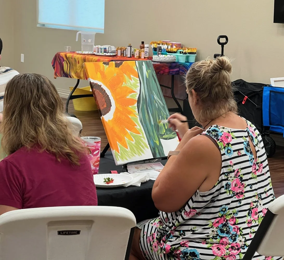 A woman painting a sunflower on a canvas during a paint and sip class, with another person sitting nearby observing.