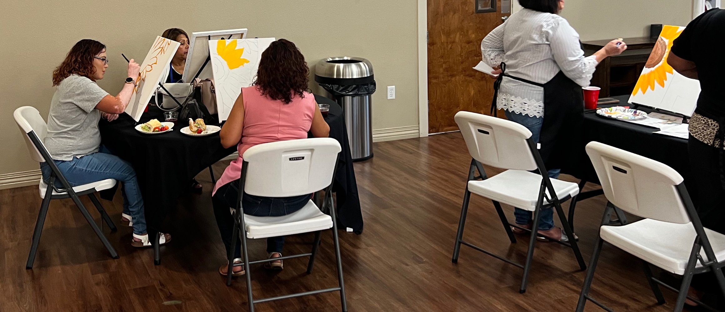 Women painting on canvases at a social gathering, with plates of food on the table in front of them and a trash can nearby.