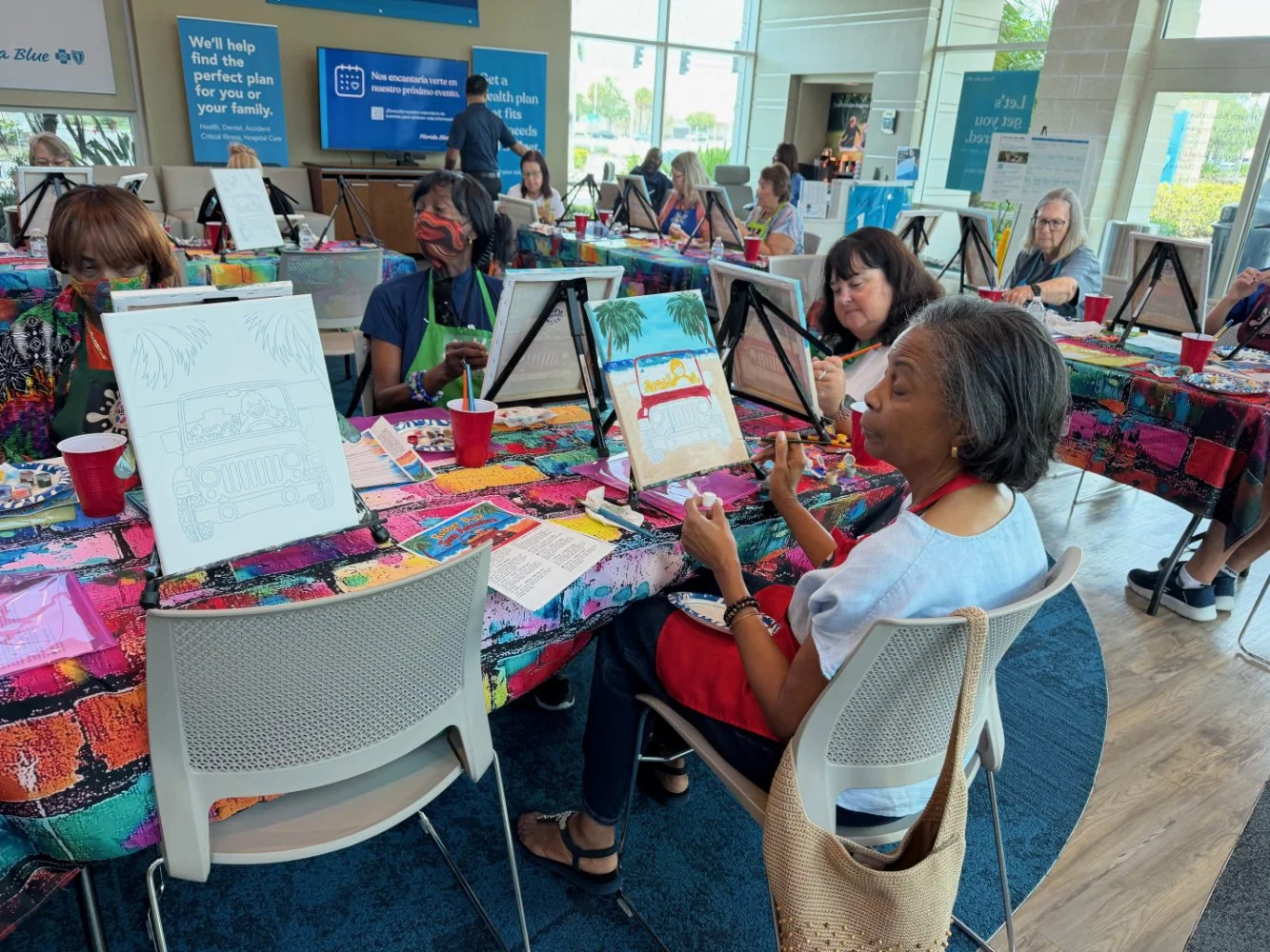 A group of people participating in a painting class at a community center or gallery. They are seated at tables covered with colorful, paint-splattered tablecloths, working on canvas paintings of a beach scene with palm trees and a car with a surfboa
