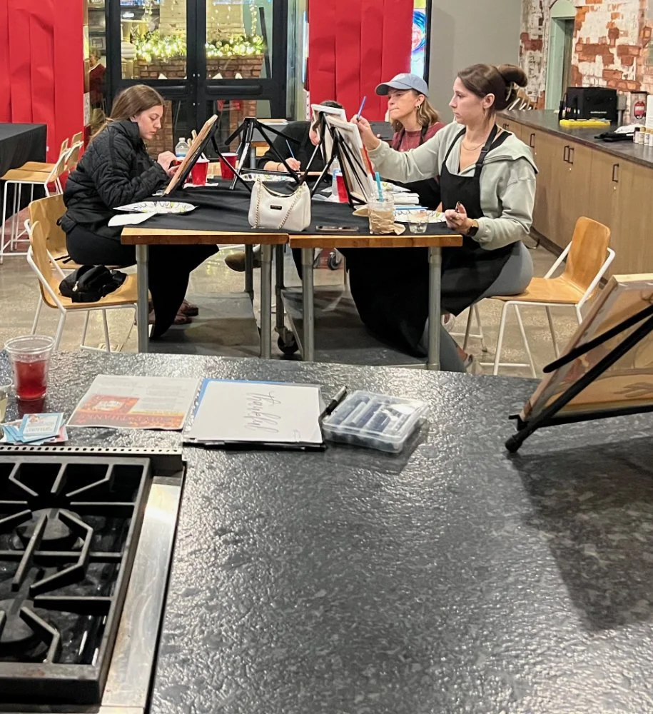 Three women painting on canvases at a table inside a cafe or restaurant.