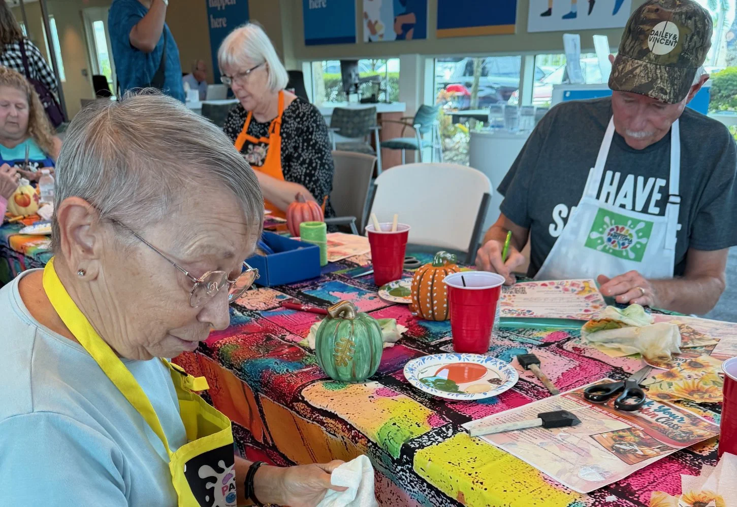 Group of elderly people participating in a craft activity at a colorful table with decorative pumpkins, scissors, and craft supplies.
