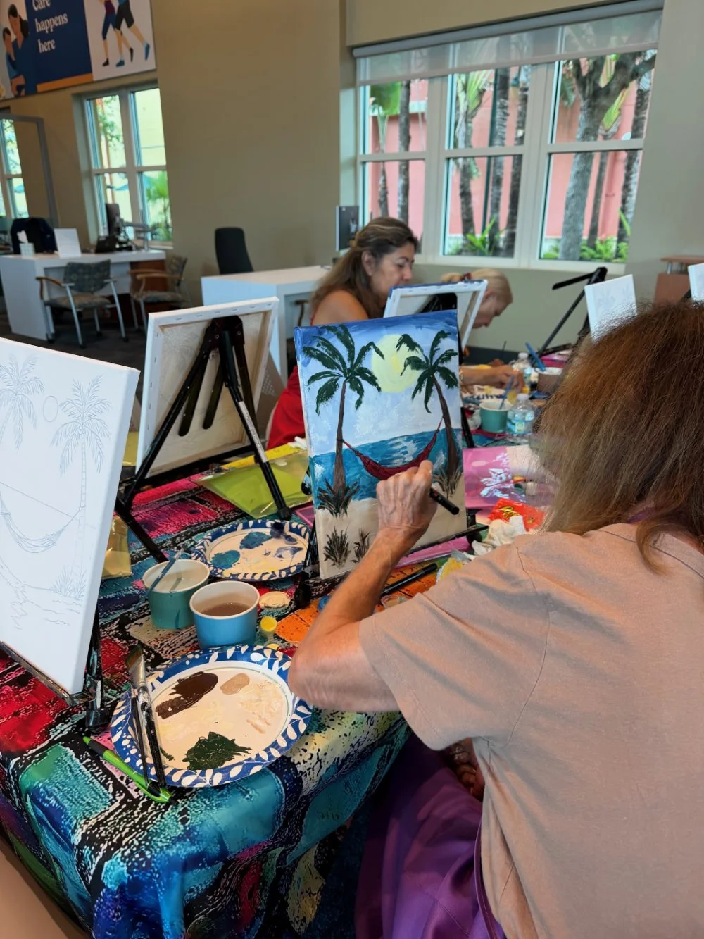 People participating in a painting class, creating a tropical beach scene with palm trees and a hammock, with art supplies on a colorful table.