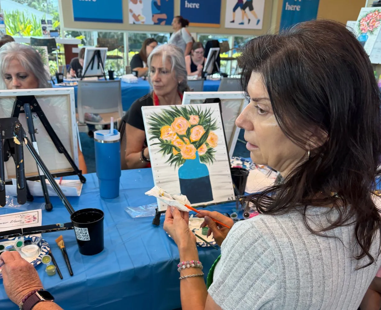 Woman painting a floral still life on a canvas at an art class with other women painting and socializing in the background.
