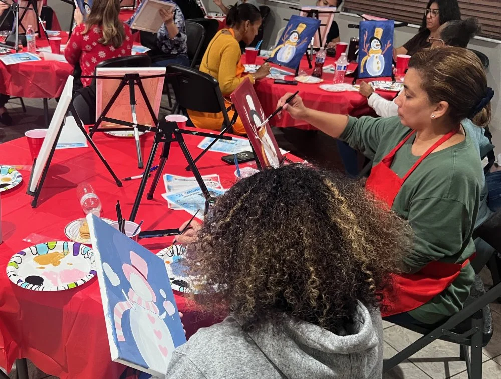 People painting snowmen on canvas at a holiday-themed group event, with red tablecloths and festive decorations.