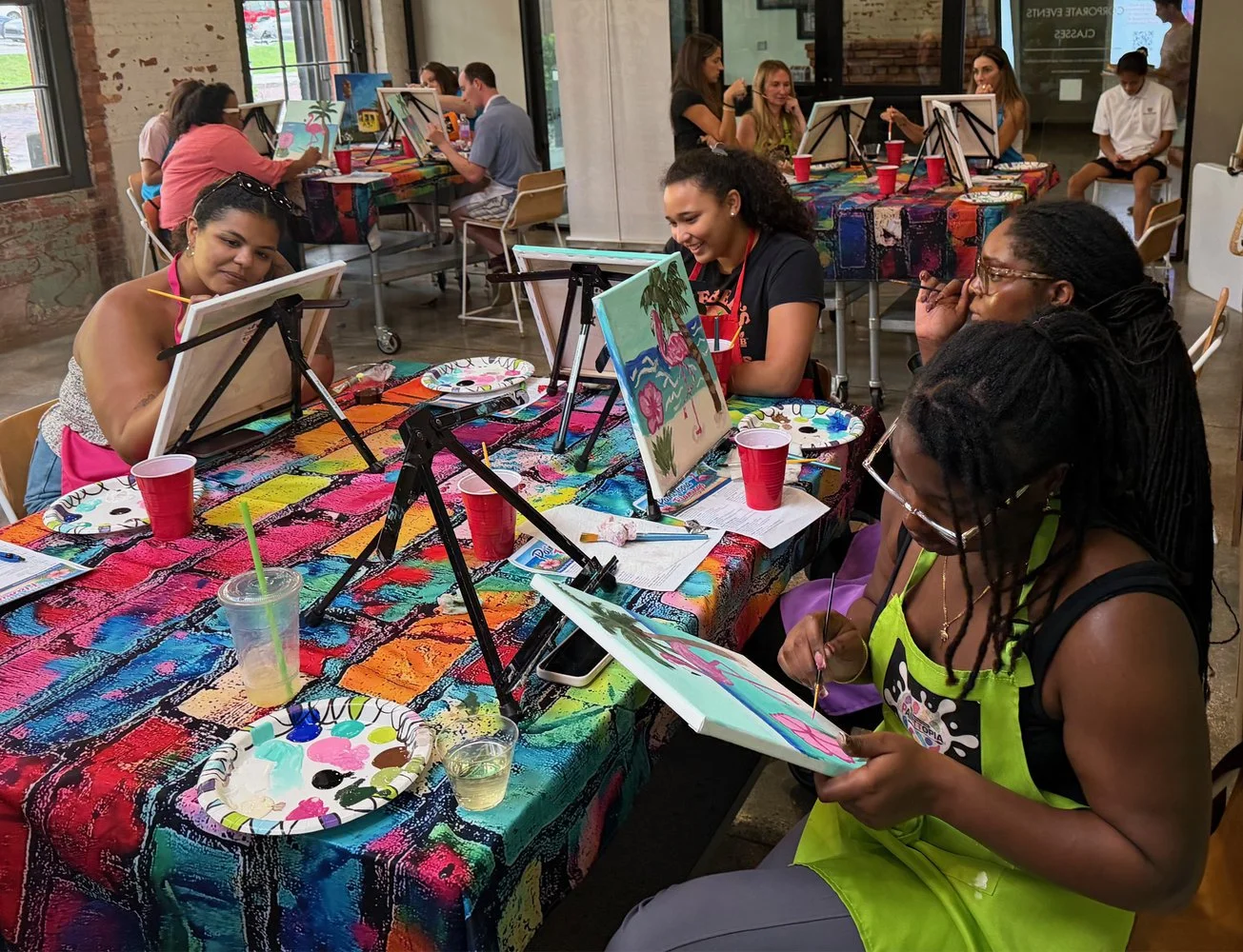 People participating in a group painting activity at a colorful table with paintings, art supplies, and drinks in a bright, industrial-style room.