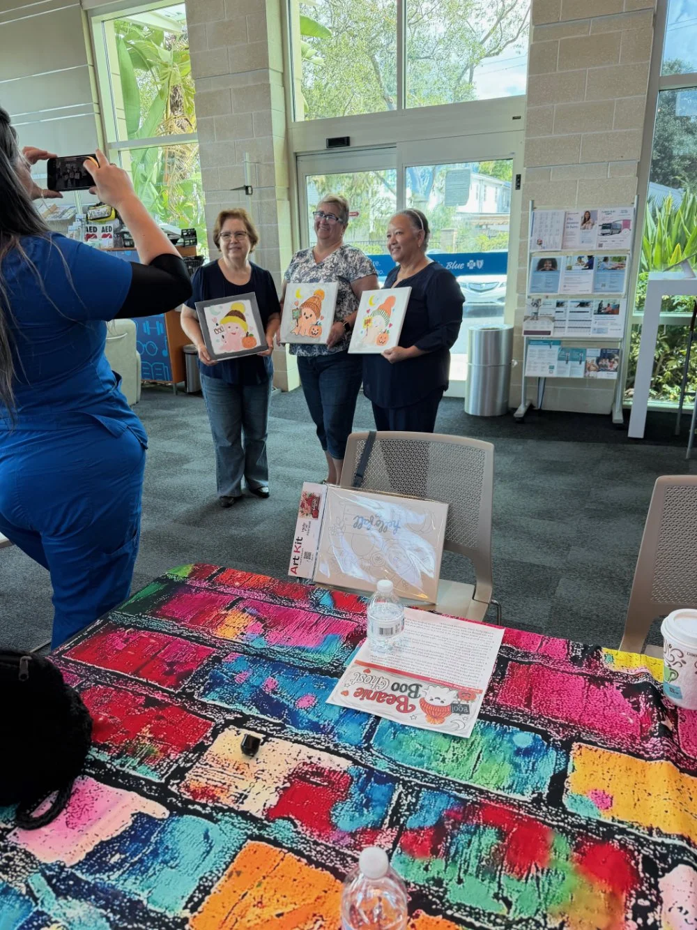 Three women are standing indoors near a large glass window, holding colorful artwork. A woman in blue scrubs is taking their photo with a smartphone. In the foreground, a table is covered with vibrant art supplies, and some printed materials, includi