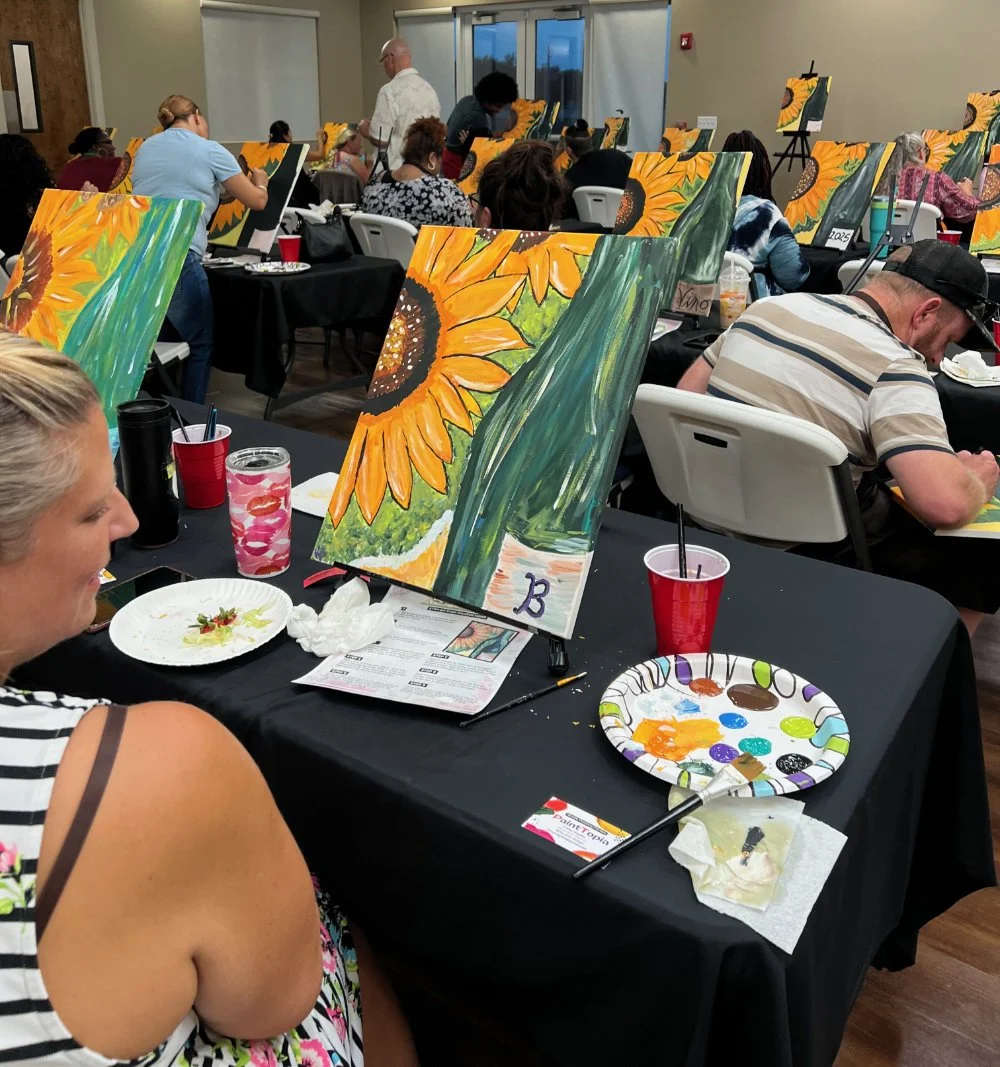 Participants painting sunflower artwork at a group art class, with paintings on easels and art supplies on tables.
