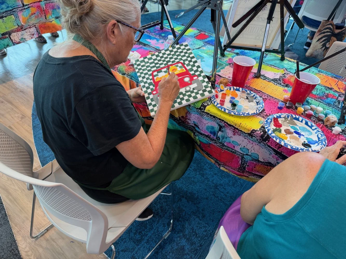 An elderly woman painting a red pickup truck on a canvas at a colorful art table with paint palettes, cups, and brushes.