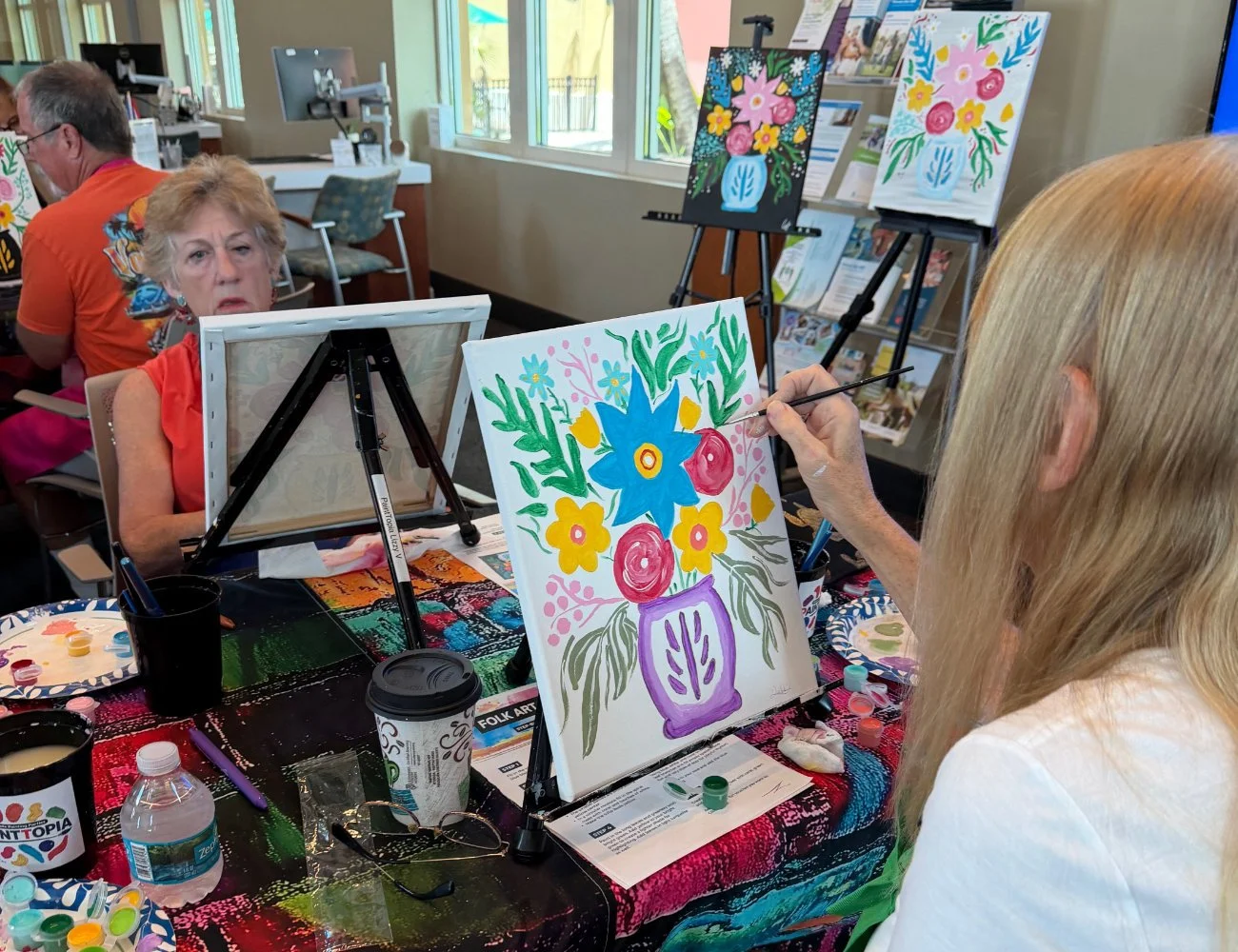 Woman painting a colorful floral still life on canvas, seated at a table with painting supplies in a group art class.