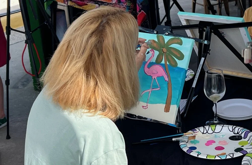 A woman with blonde hair is painting a picture of a pink flamingo standing next to a palm tree on a canvas at a painting party.