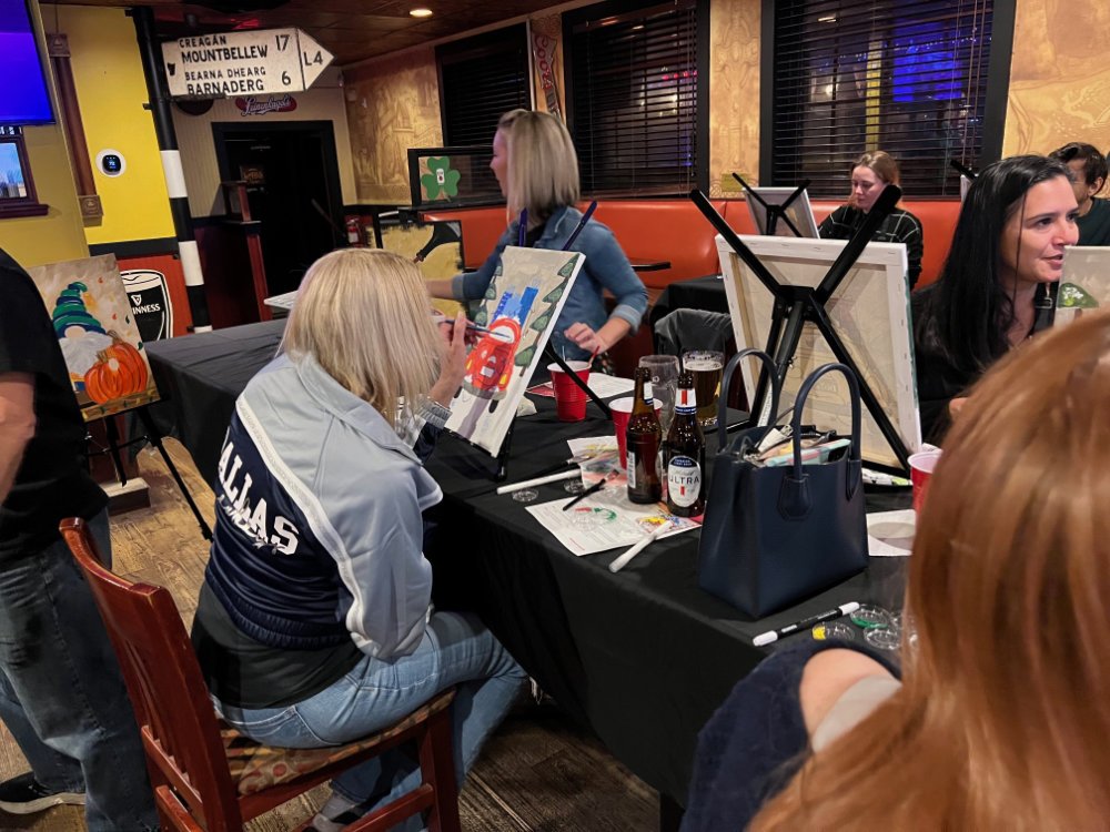 People participating in a painting class at a restaurant or bar, with paintings of pumpkins and other seasonal themes on easels.
