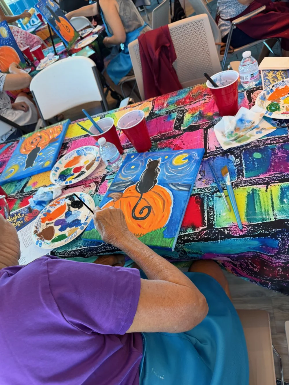 Older person painting a picture of a black cat sitting on an orange pumpkin against a starry night sky on a colorful tablecloth.