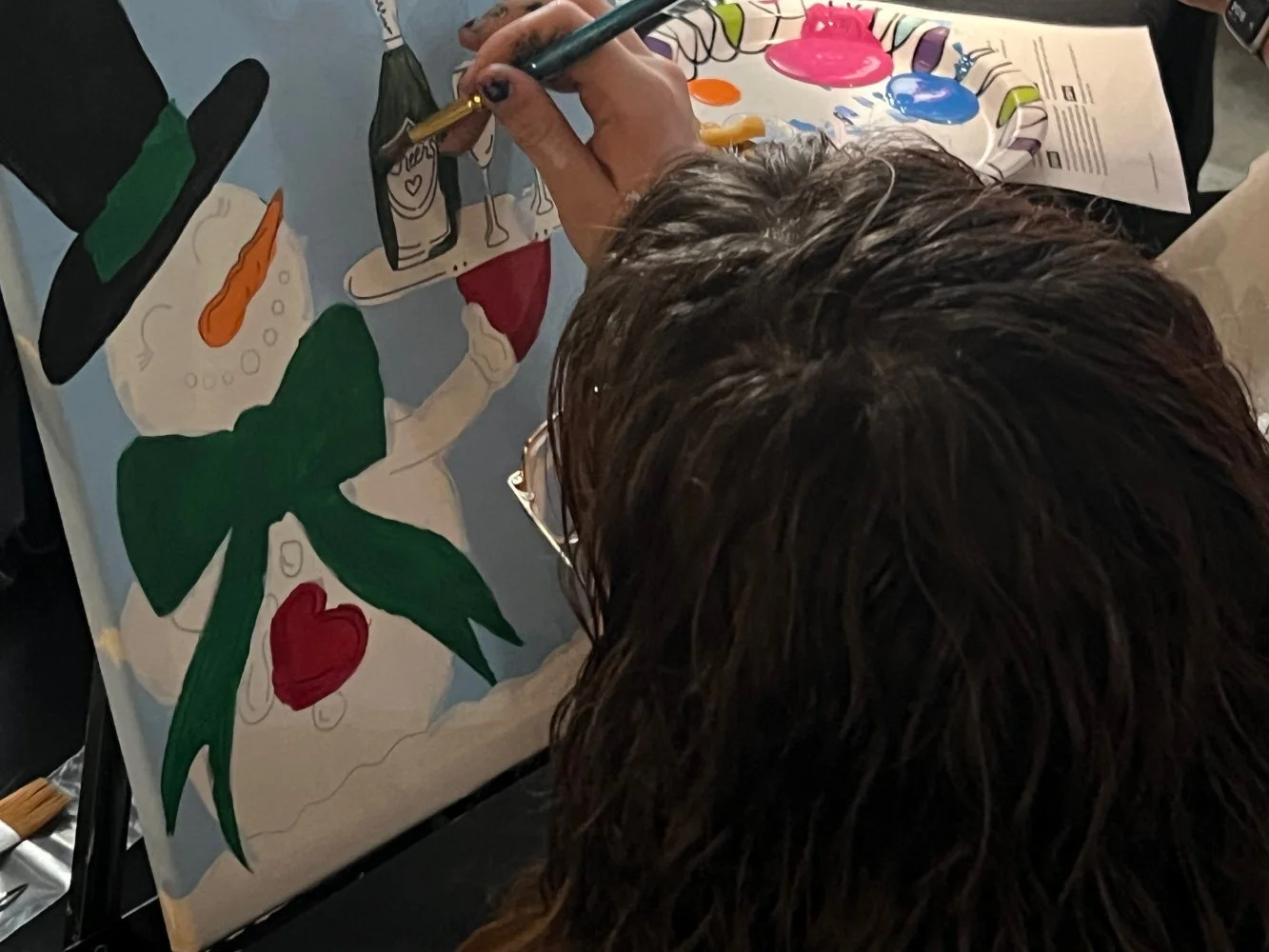 A person with brown hair is painting a colorful picture of a snowman decorated with a green bow, a red heart, and a black hat, using various paints and brushes on a canvas.