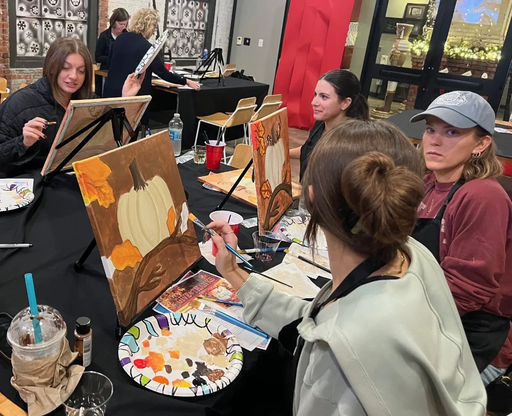 People painting fall-themed scenes with pumpkins and autumn leaves on canvases at a group art class.