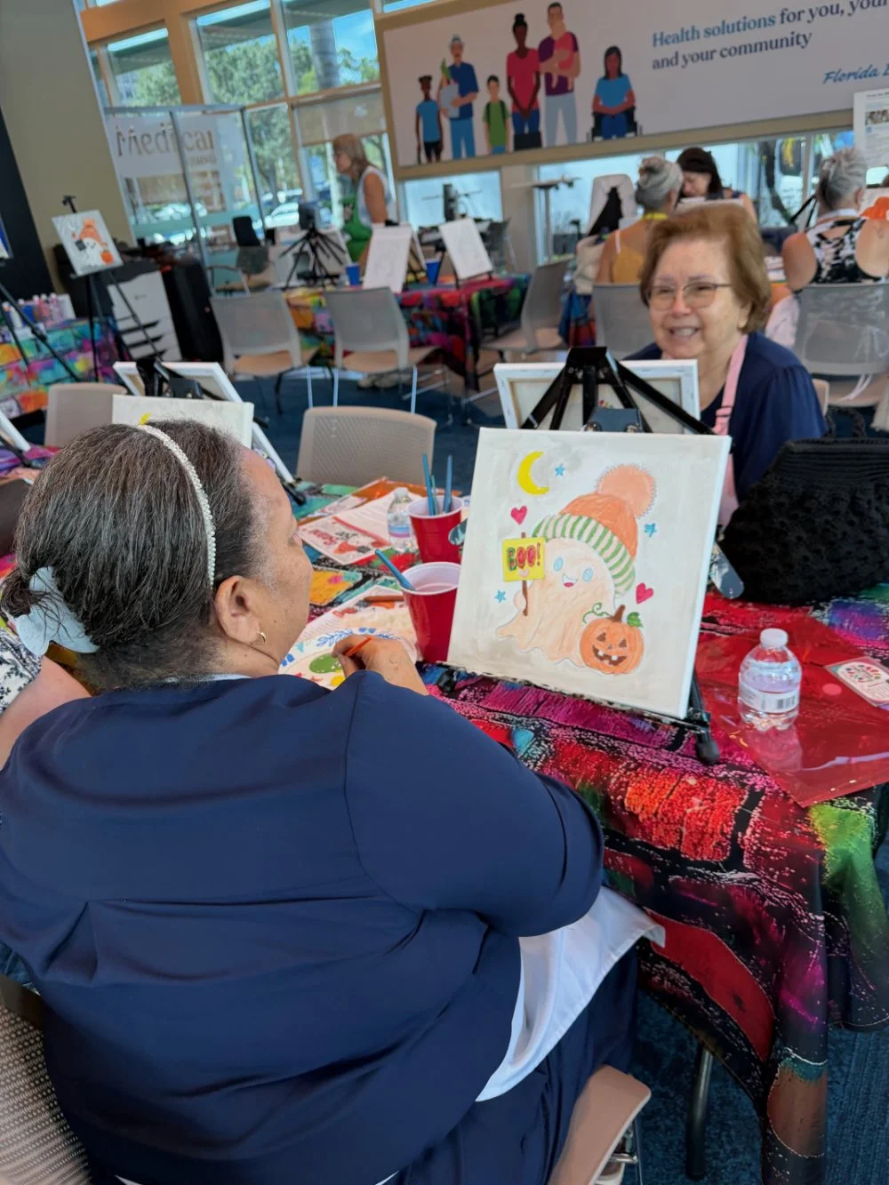 Women painting Halloween-themed picture at an art class, with smiling woman in background, colorful tablecloth, painting supplies, and art-themed posters in the background.