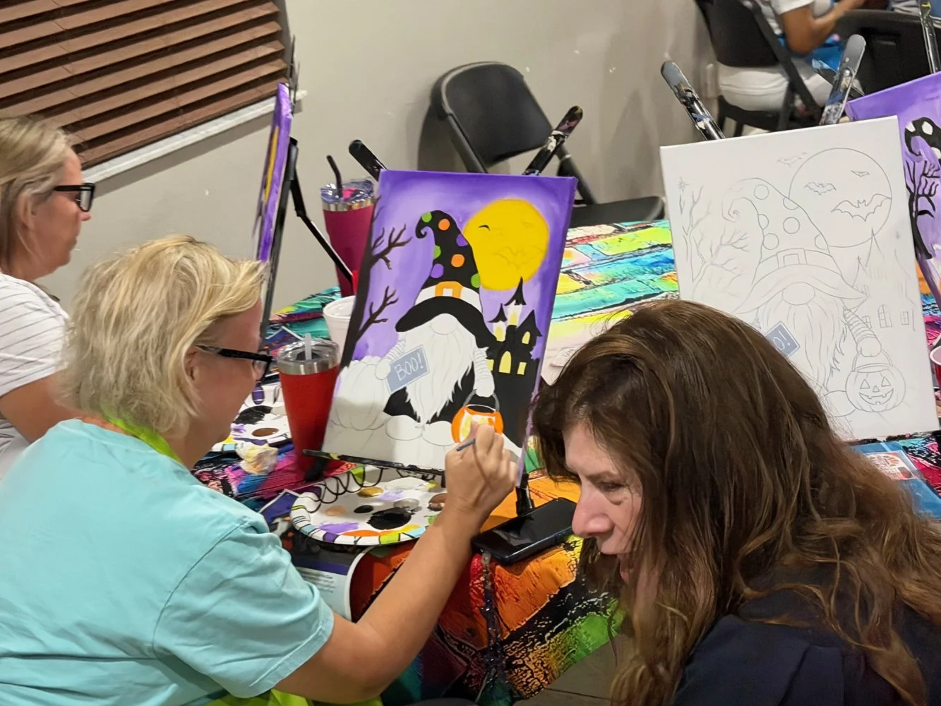 People sitting at a table painting Halloween-themed pictures, with one canvas showing a witch with a polka-dot hat, a haunted house, and a bright yellow moon.