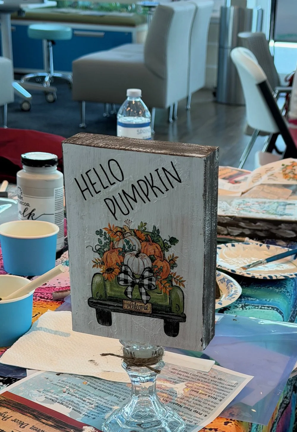 Decorative sign with pumpkins and fall-themed floral arrangement, reading "Hello Pumpkin" and "Welcome" on a table with craft supplies and paper plates, in a bright room with chairs and a water bottle in the background.