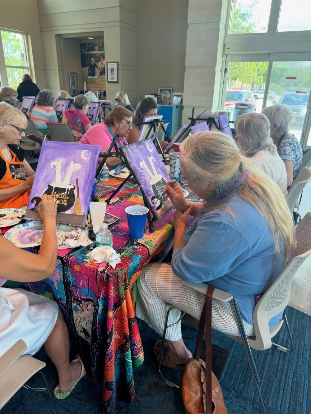 A group of older women participating in a painting class, working on Halloween-themed paintings featuring a bubbling cauldron labeled 'Hocus Pocus' with a purple background, sitting at a colorful table inside a well-lit room with large windows.