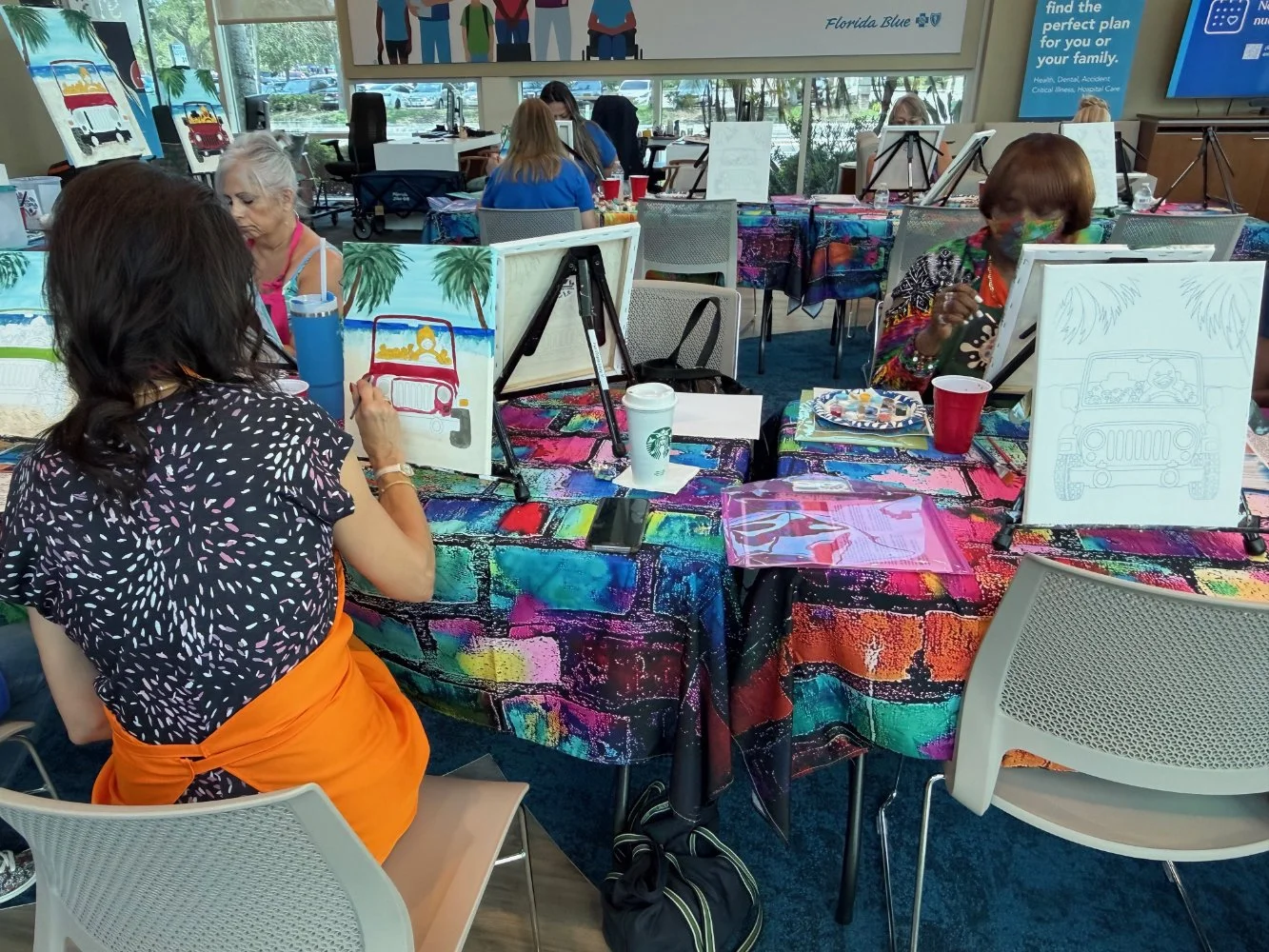 Women participating in a painting class at a colorful table with paintings of tropical scenes, some women wearing masks, in a bright room with large windows.