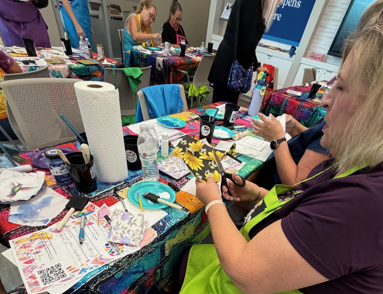 Women participating in a craft workshop at colorful tables. Supplies like paper towels, paintbrushes, bottles of water, and decorative paper are visible on the table.