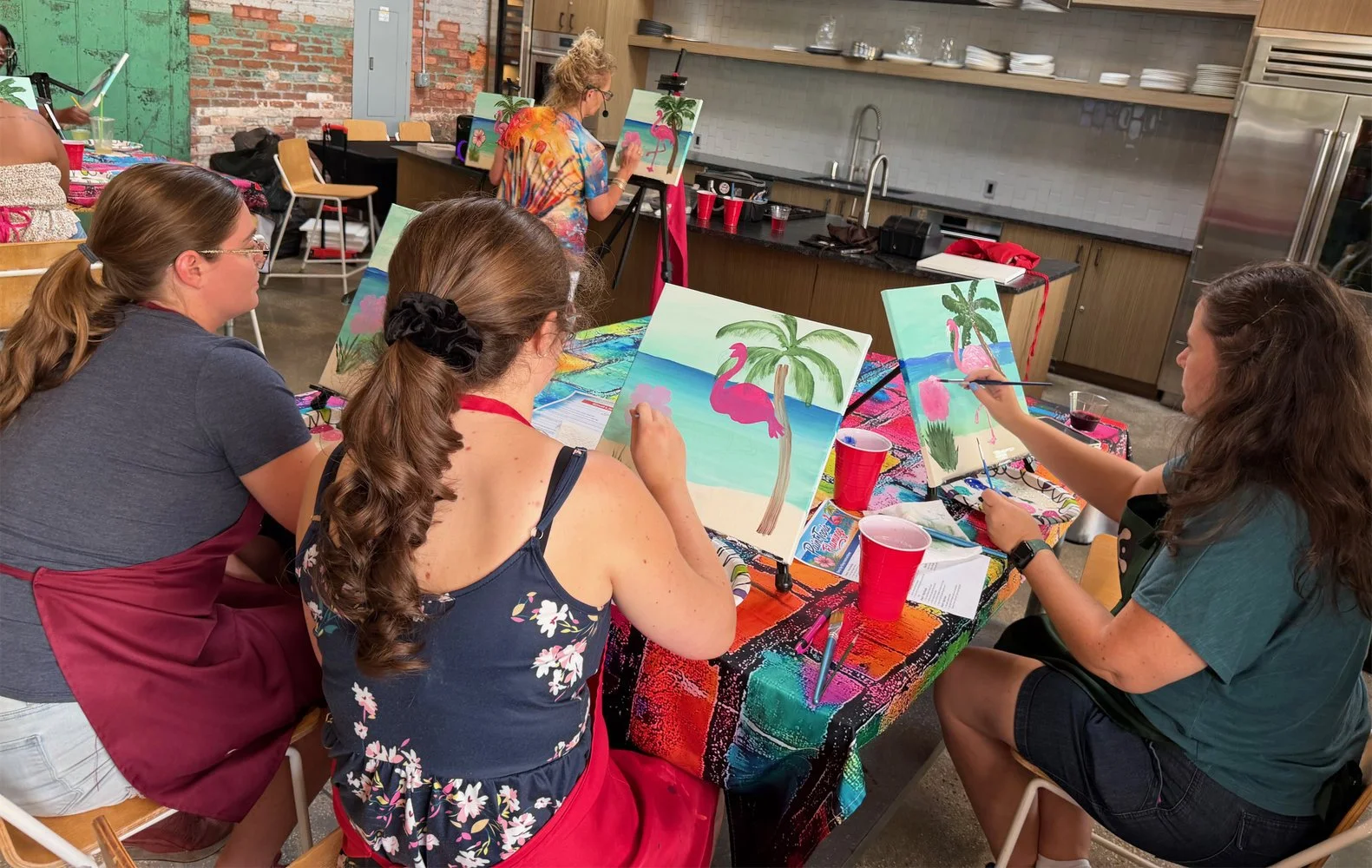 People participating in a group painting class in an indoor setting with exposed brick and wood shelves, painting tropical scenes with pink flamingos on canvases, drinks on the table, and an instructor in the background.