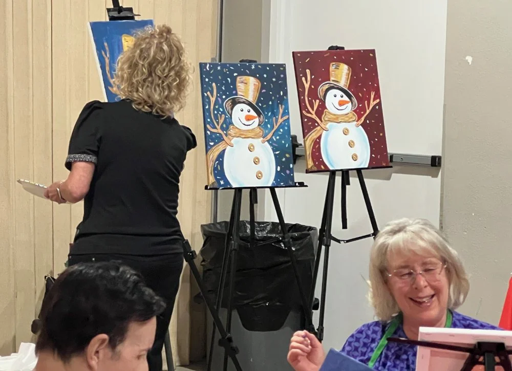 A woman painting snowmen on canvases at an art event, with finished snowmen on display, and laughing women nearby.