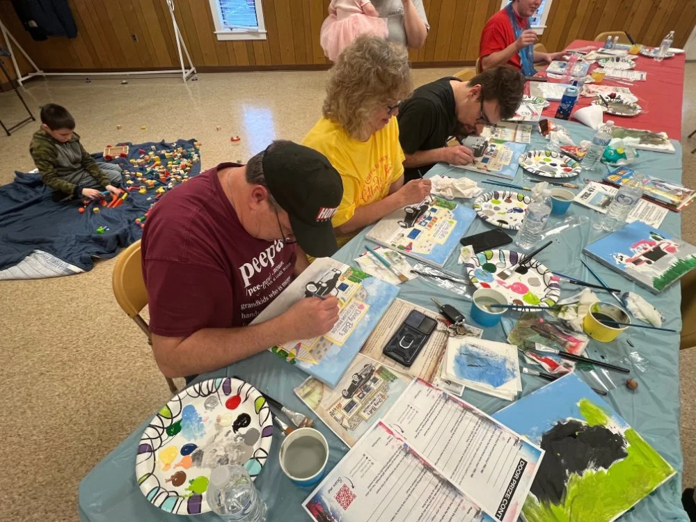 People sitting at a table painting on canvases with a variety of paints and brushes, while a child plays with building blocks on the floor.