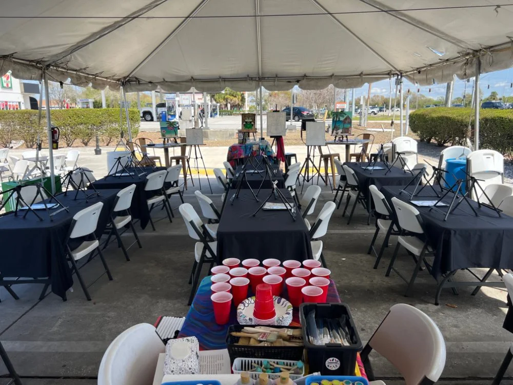 Party setup under a large white canopy tent with tables covered in black tablecloths and white chairs. A table with red cups, napkins, and utensils is in the foreground. In the background, there are easels with paintings and a parking lot.
