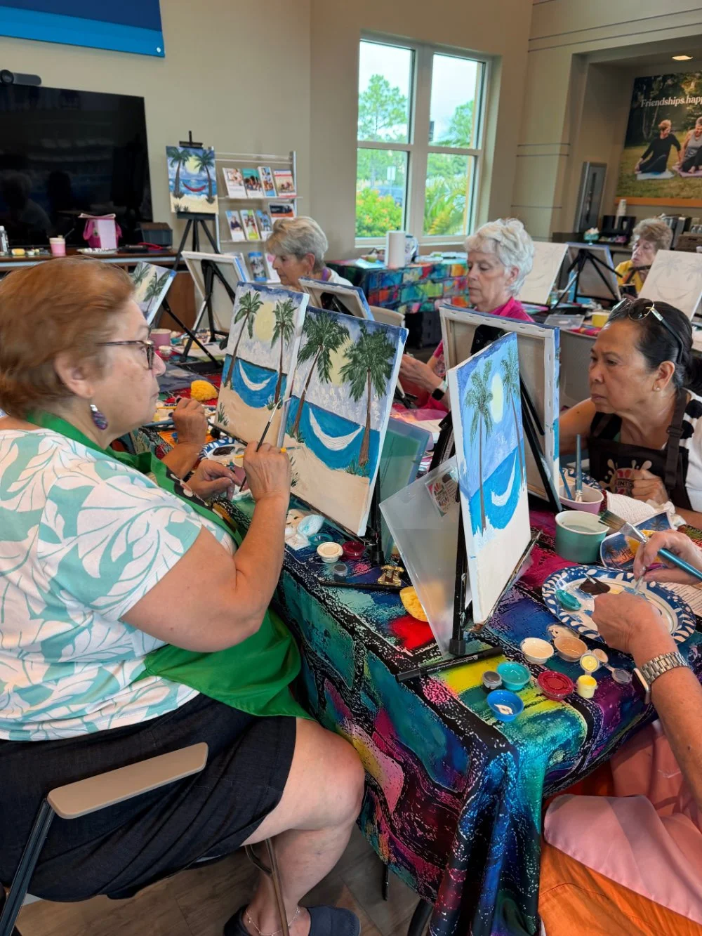 Group of older women painting tropical beach scenes with palm trees and hammocks in an art class, sitting at a colorful table inside a room with windows.