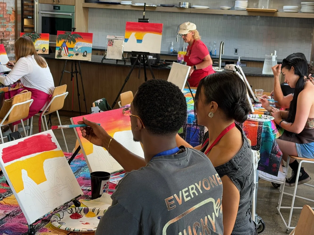 People participating in a group painting class, with canvases, paints, and brushes, some partially painted with sunset scenes and vehicles, in a modern indoor studio.