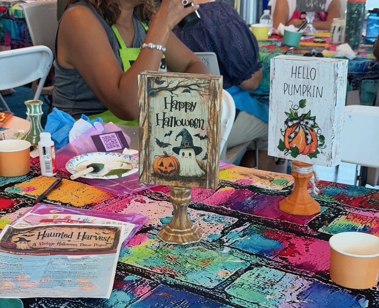 Two Halloween-themed signs on a colorful tablecloth at a Halloween craft or decor workshop. One sign says "Happy Halloween" with a ghost, pumpkin, and bats, while the other says "Hello Pumpkin" with a pumpkin illustration. Various craft supplies and 