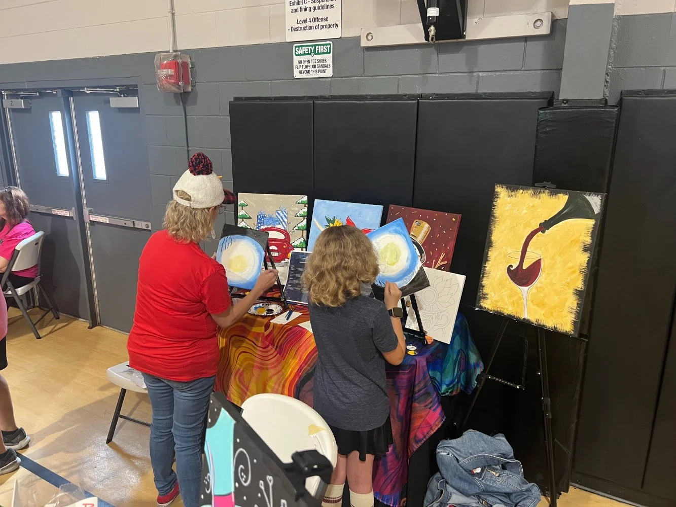Two children painting at an indoor art event, with paintings of eggs, holiday scenes, and a wine glass on display, against a gymnasium wall.
