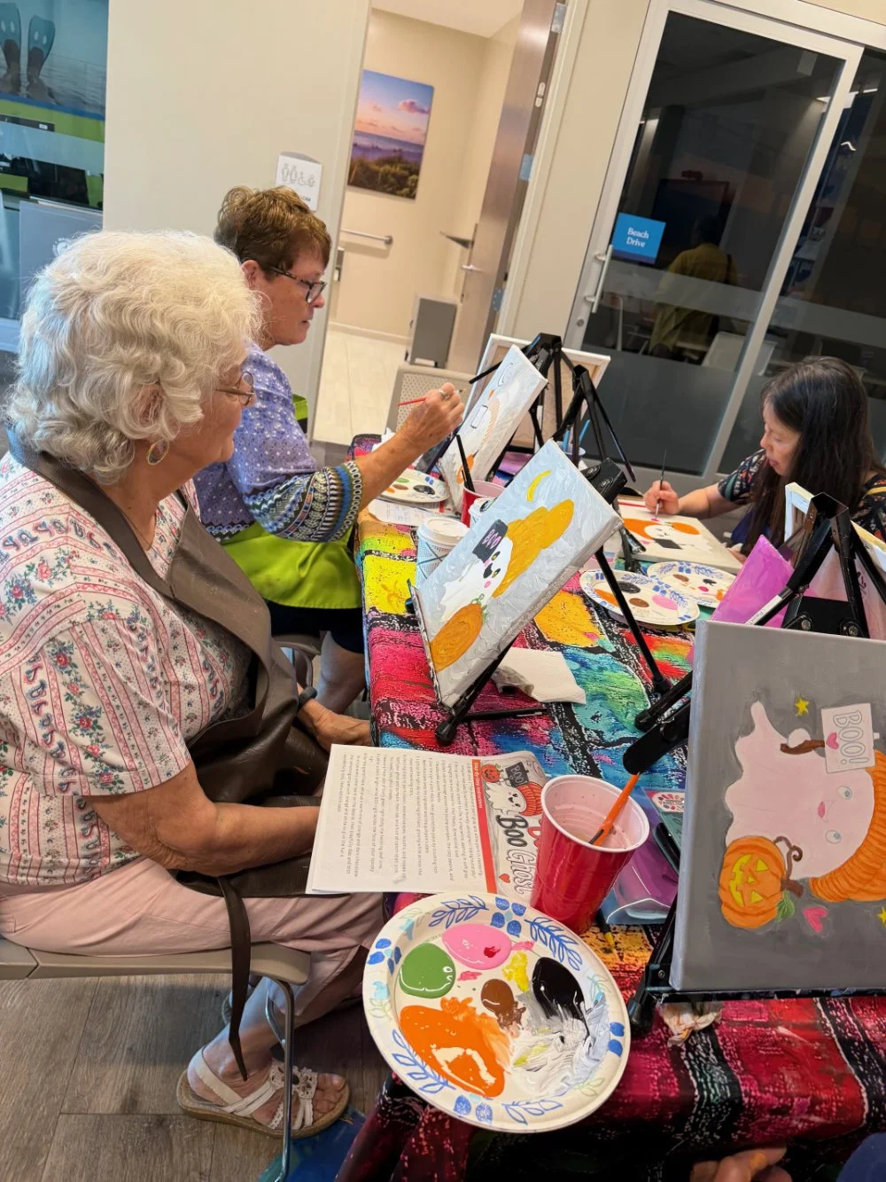 Four women sitting at a table painting Halloween-themed canvases inside a room. The table is covered with a colorful cloth, and painting supplies are visible. One woman in the foreground has white curly hair and is looking at a paper. One woman in th