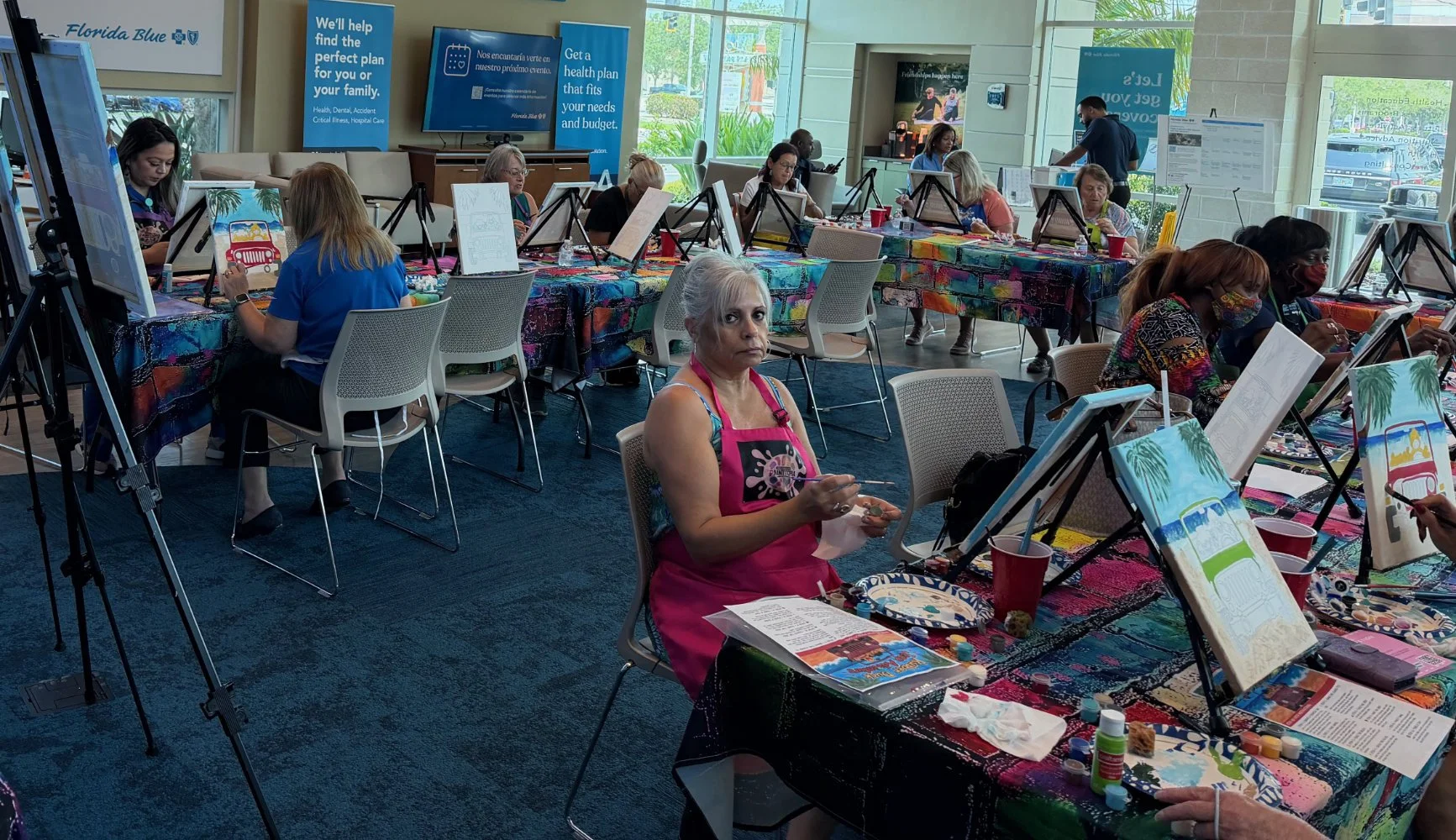 People attending a painting class in a bright room with large windows, seated at tables covered with a colorful, paint-splattered tablecloth, with canvases and painting supplies in front of them.