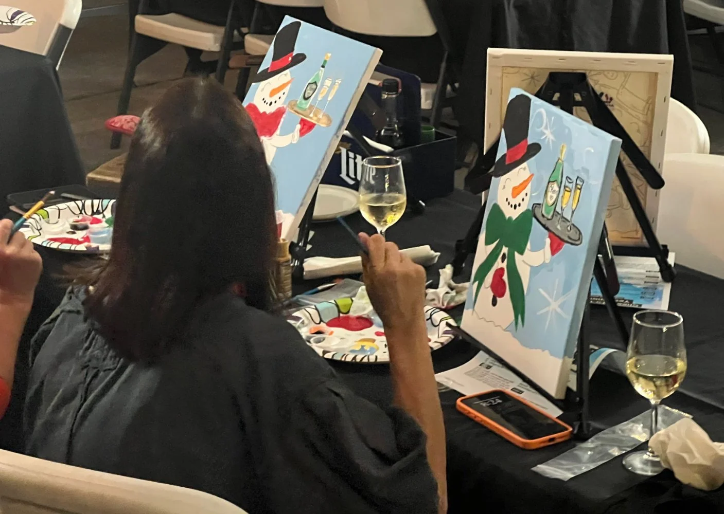 Person painting Christmas snowmen on canvases at a table with wine glasses and painting supplies.