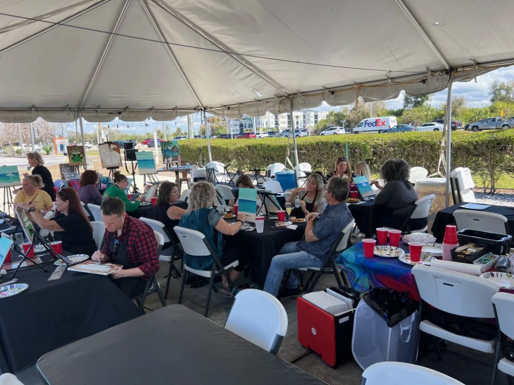 People sitting under a large tent, engaged in activities like reading, working on laptops, and socializing at tables during daytime, with a hedge and parked cars in the background.