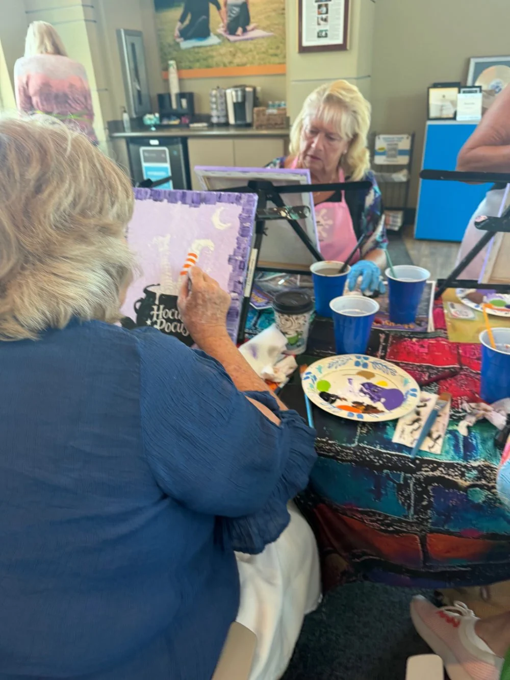 Women painting canvas boards at a table with paint supplies and drinks.