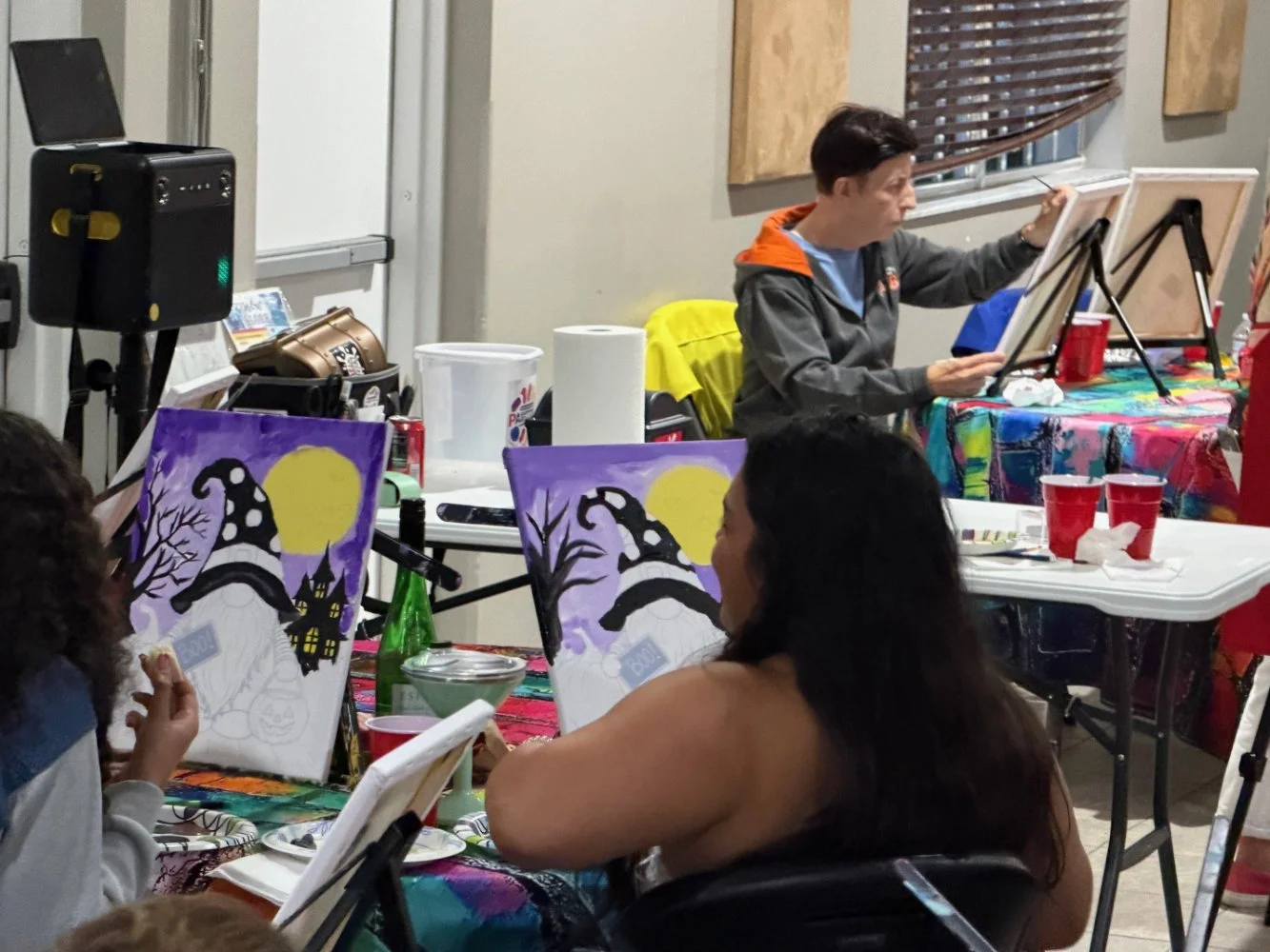 People painting Halloween-themed pictures of witches and haunted houses at an indoor painting party.