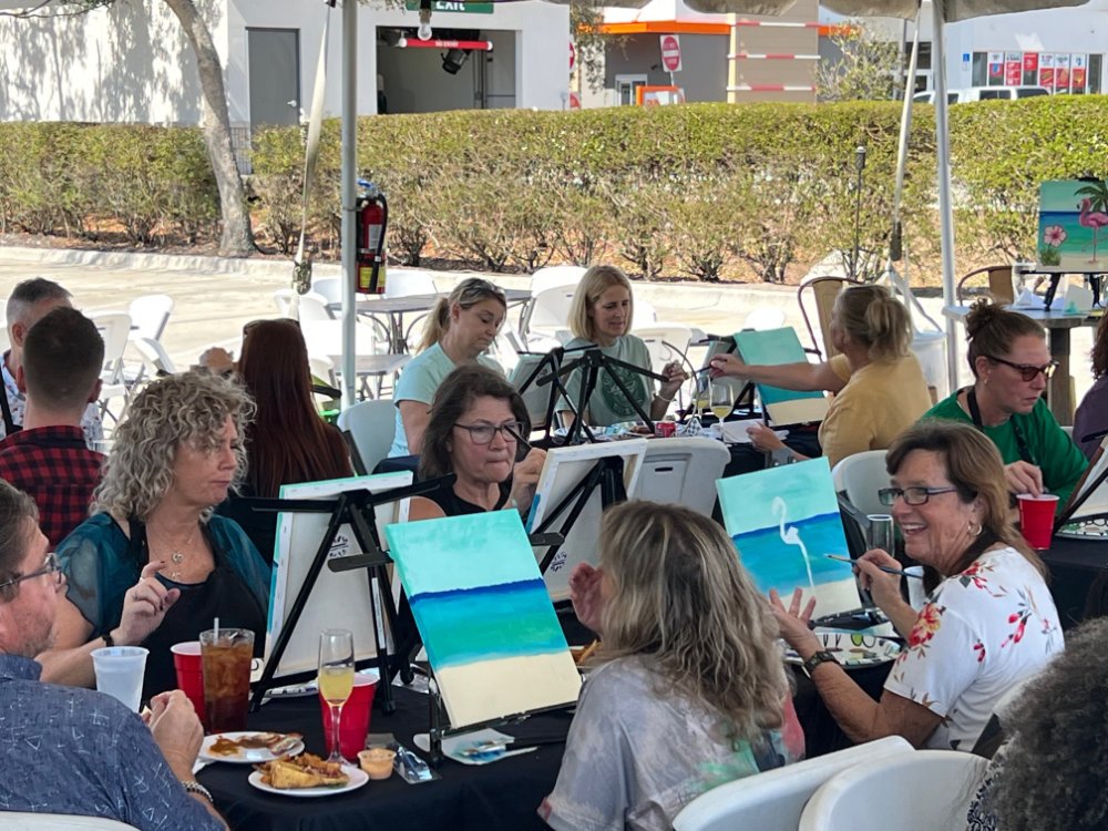 A group of people participating in a paint-and-sip event outdoors under a canopy. They are painting seascapes, featuring blue waters and white herons, and are seated at tables with supplies and beverages.