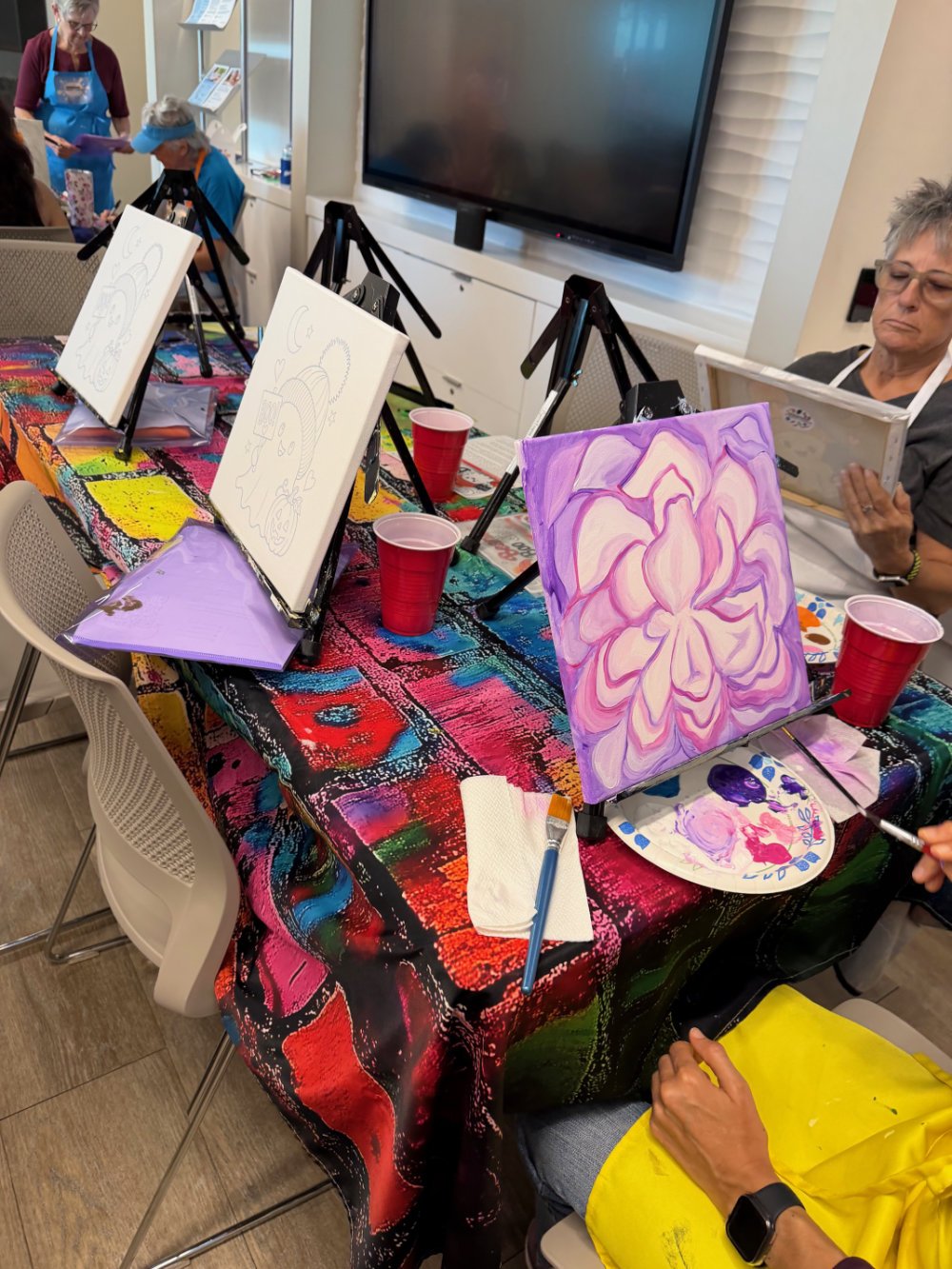 People participating in a group painting activity at a table covered with a colorful tablecloth. Several canvases with sketches and painted designs are propped on small easels. Painting supplies, cups, a palette, and a paper towel are on the table.