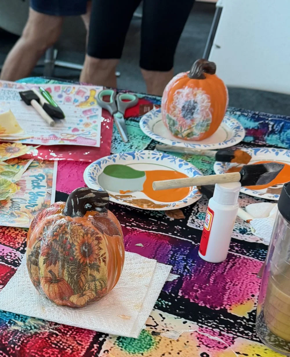 Decorated craft pumpkins on colorful table with scissors, glue, paper, paint, and art supplies.