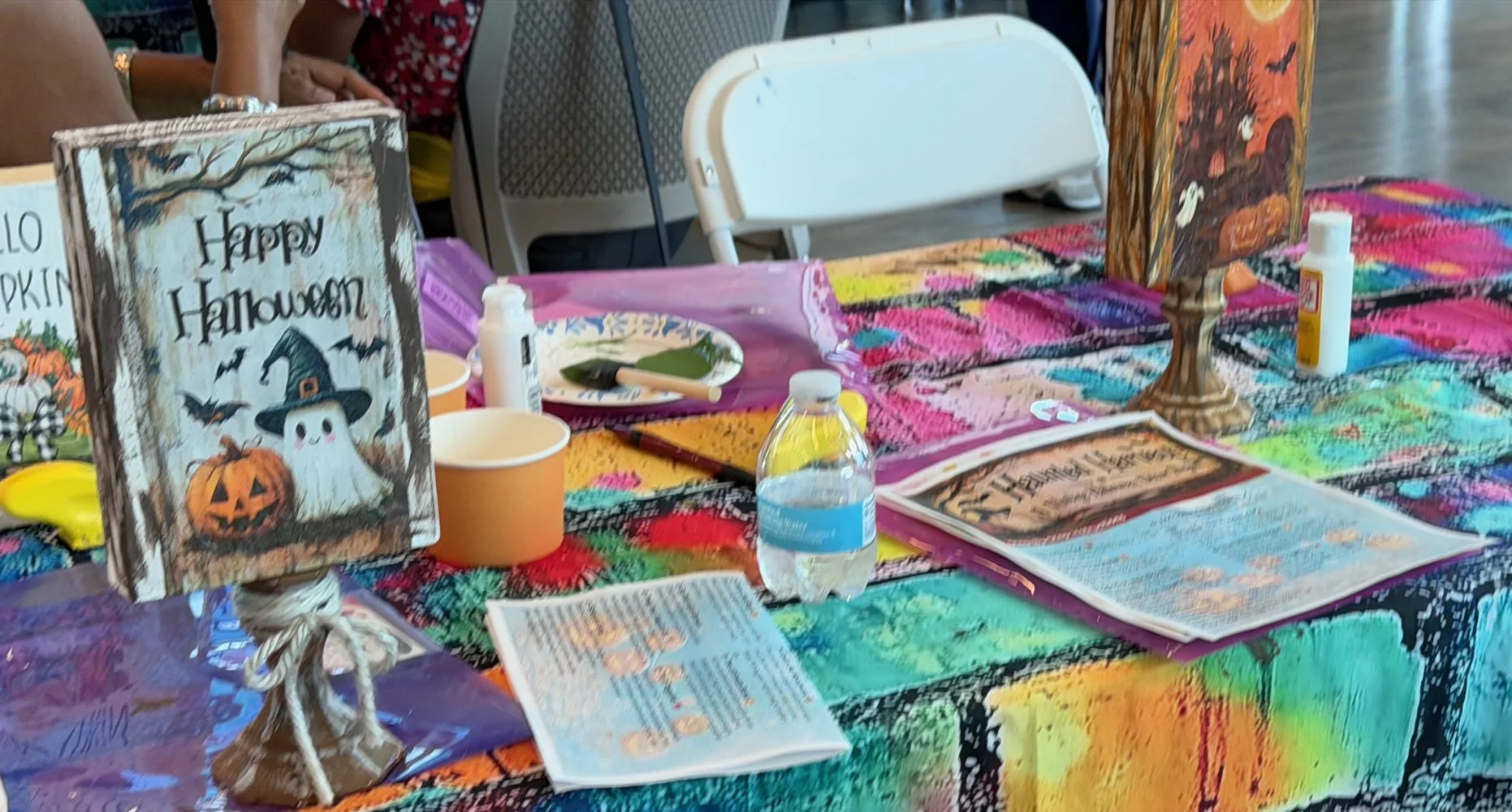 A table decorated for Halloween with colorful tablecloth, signs, and craft supplies, including a sign that reads 'Happy Halloween' with a ghost and pumpkin illustration.