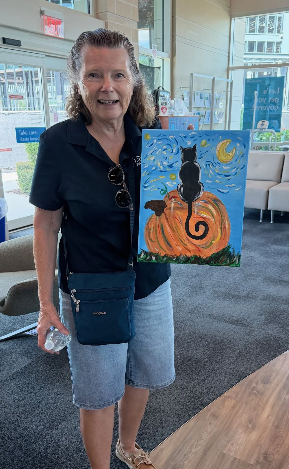 A woman with short brown hair wearing a black shirt and denim shorts is standing indoors, holding a painting of a black cat sitting on a large pumpkin against a night sky with a moon and stars.