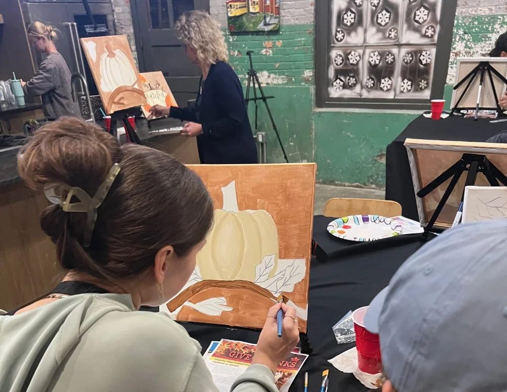 People painting pumpkins on canvases at a social art event, with some paintings in progress and art supplies visible.
